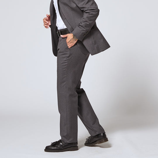 A profile shot of an androgynous model wearing charcoal dress pants, hand inside the pocket pictured as the matching blazer is swooped back
