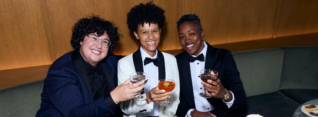 Three people in formal evening attire sitting together at a table with drinks, plates, and disco-ball style decor in an indoor lounge or event space.
