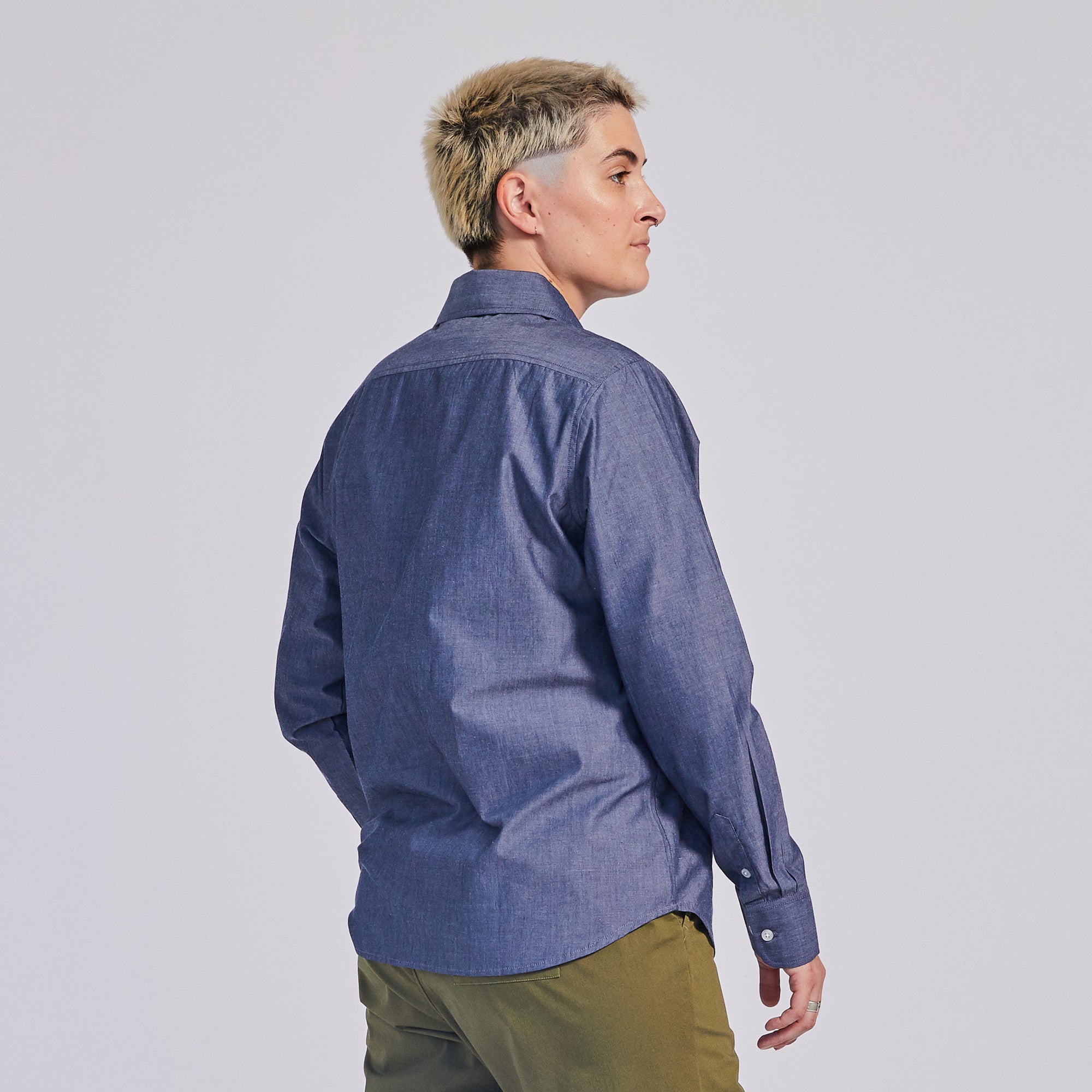 Model wearing a blue chambray long-sleeve shirt facing the wall to show off the back of the shirt. Color: Blue Chambray