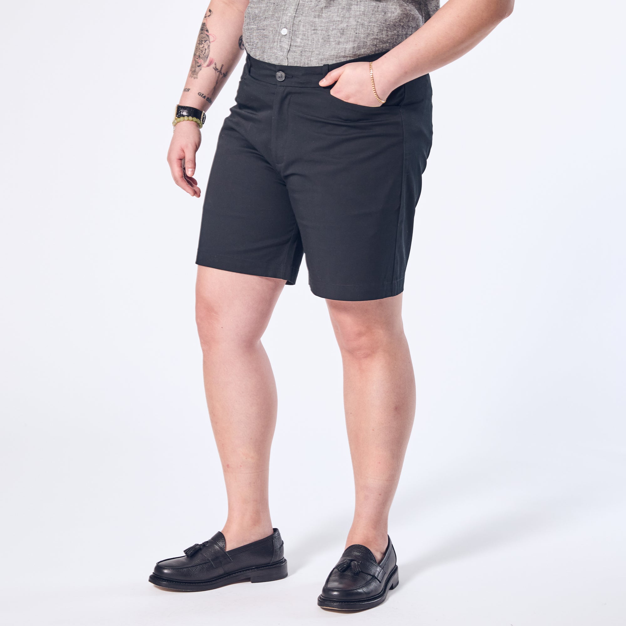 A person standing, wearing black chino shorts and black shoes, with a grey shirt and a watch on the left wrist.