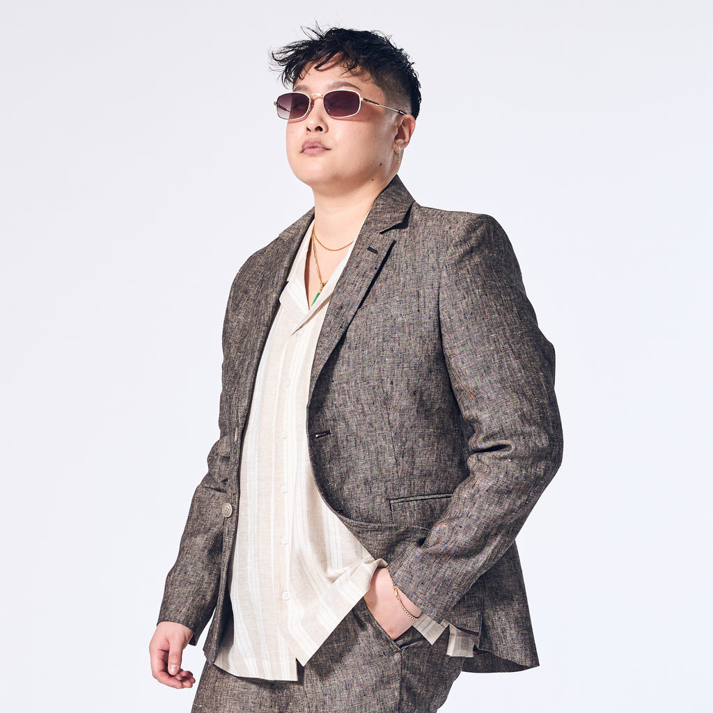 A person wearing a driftwood linen blazer with a sharp silhouette, paired with a light colored shirt and sunglasses.