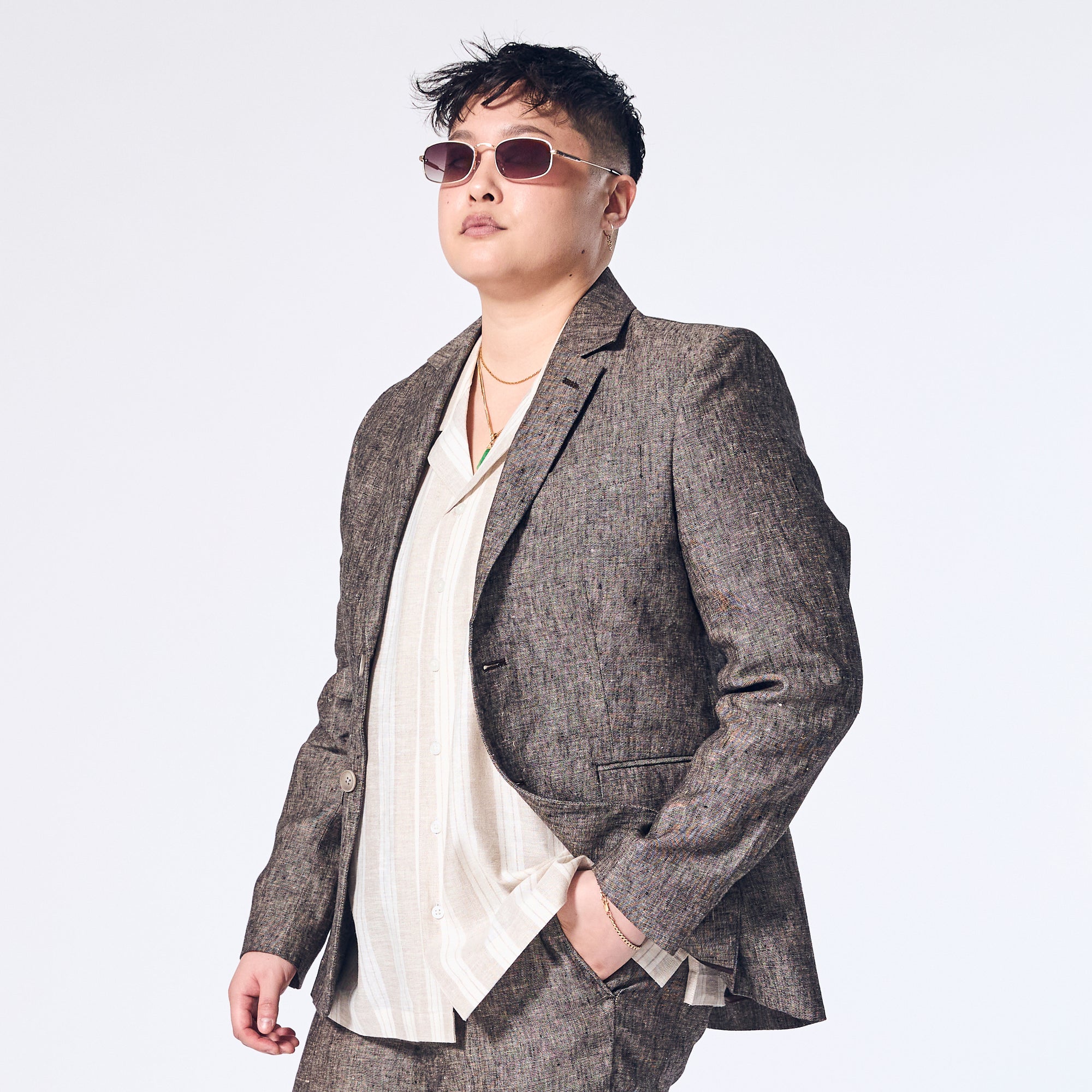 A person wearing a driftwood linen blazer with a sharp silhouette, paired with a light colored shirt and sunglasses.