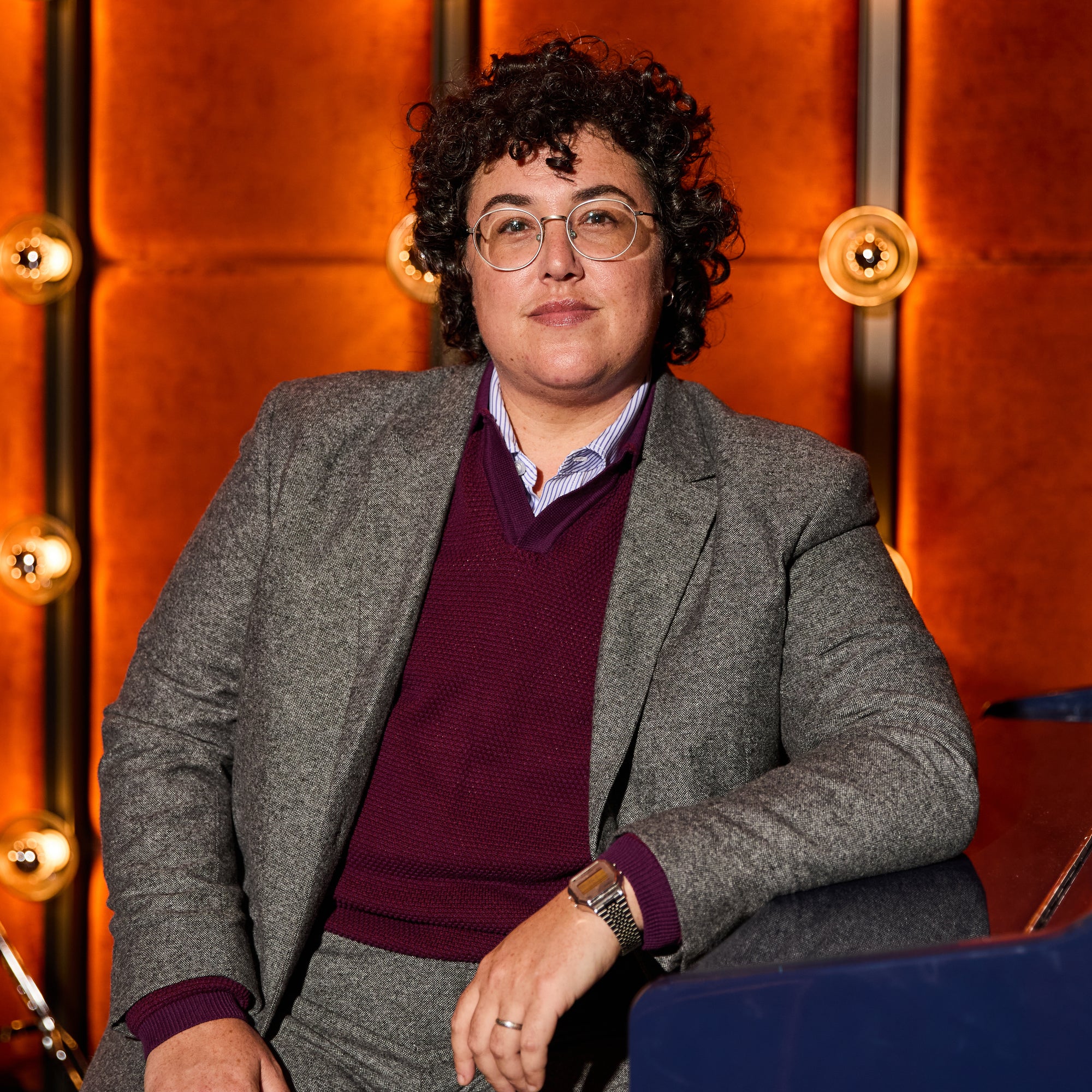 Person wearing glasses and a gray blazer with a maroon shirt against an orange wall with lights.