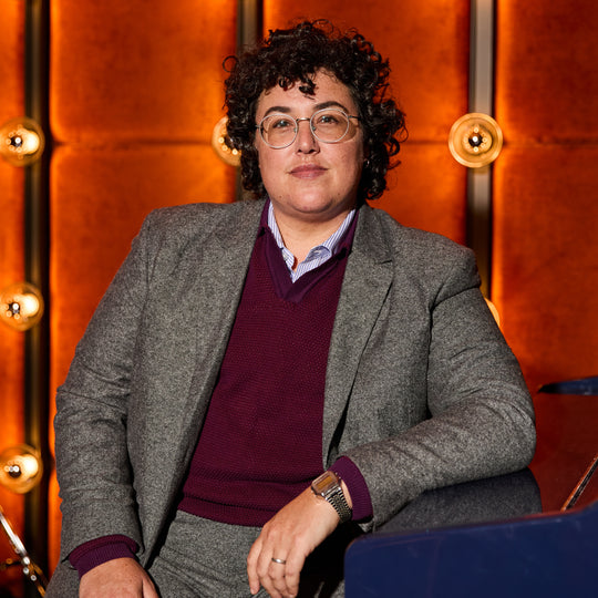 Person wearing glasses and a gray blazer with a maroon shirt against an orange wall with lights.