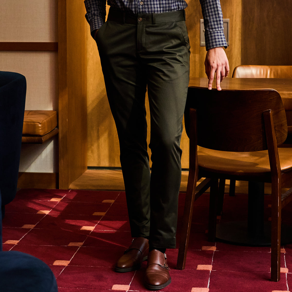 Person wearing olive chinos and brown shoes standing in a room with wooden furniture and red carpet. Color: Olive
