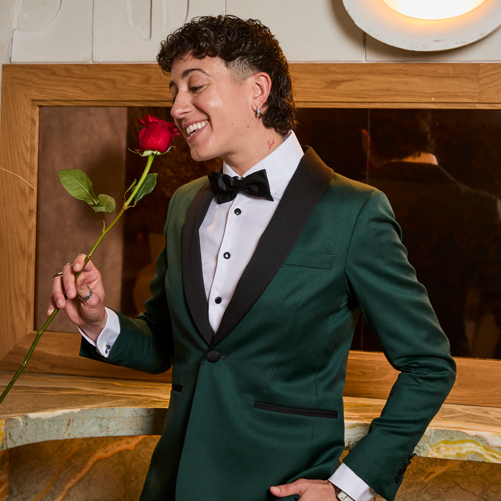 Model in a green tuxedo holding a red rose indoors. Color: Emerald