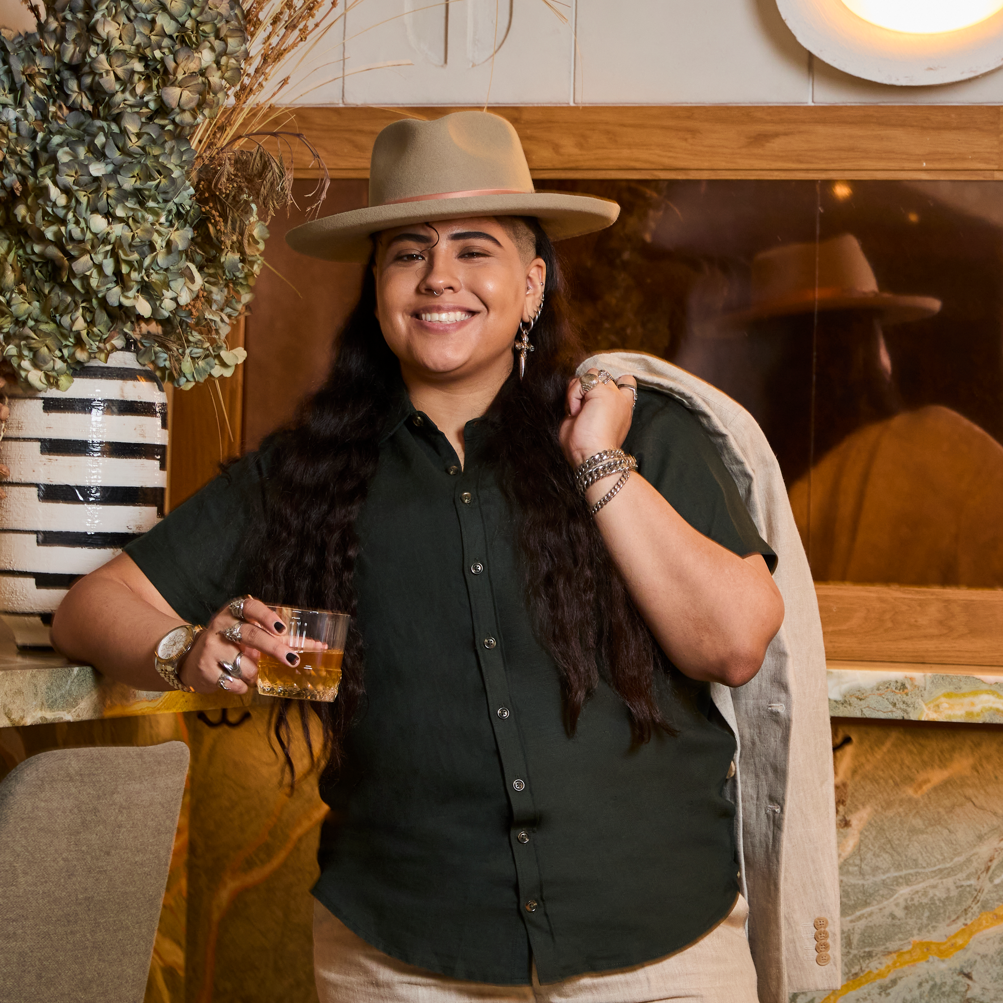 Model wearing a cowboy hat and holding a drink, standing in a cozy indoor setting. Color: Nori