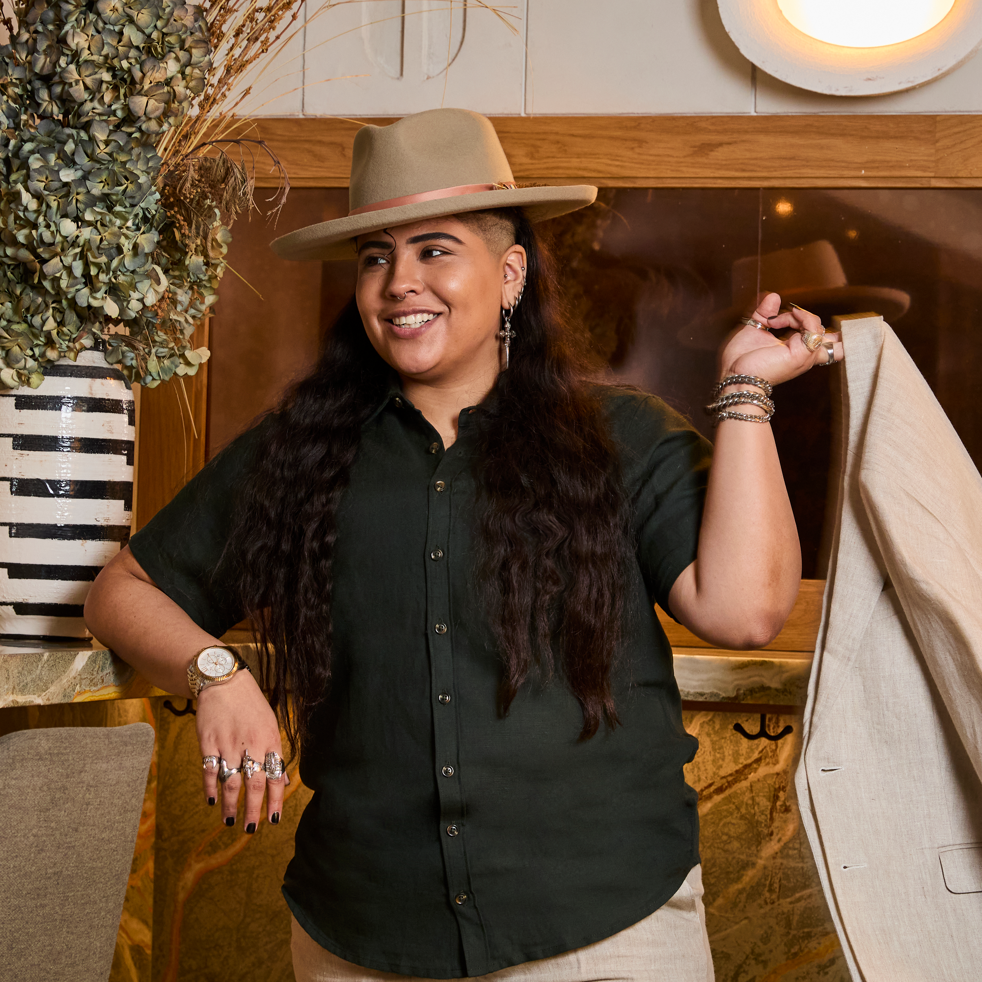 Model wearing a dark green shirt and beige hat, holding a beige jacket indoors. Color: Nori