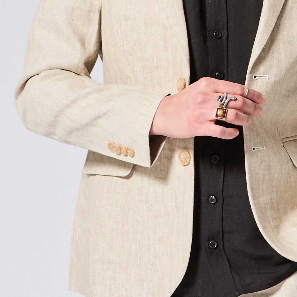 Person wearing a beige blazer with gold buttons and a black shirt, holding a small object. Color: Khaki Herringbone