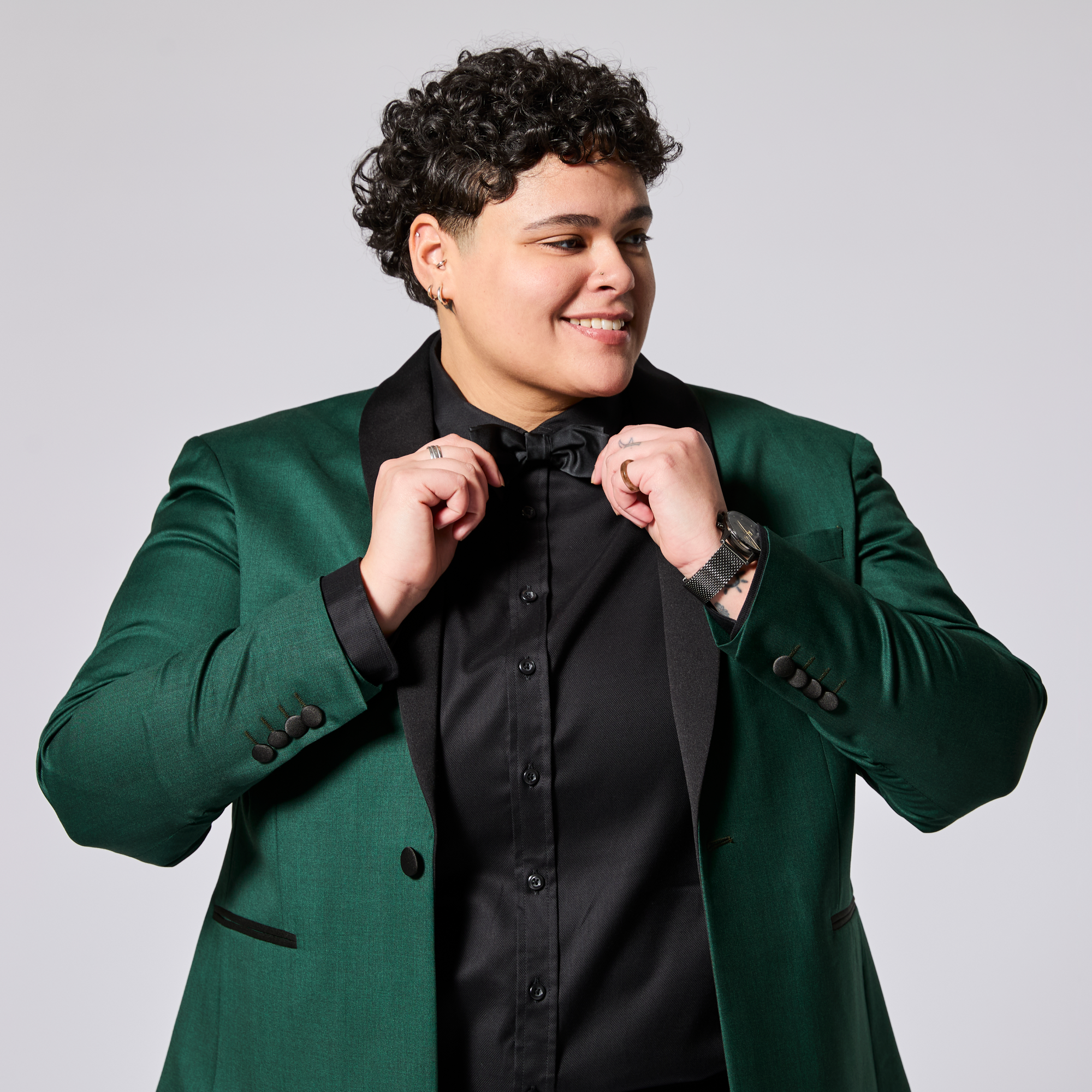 Model in emerald tuxedo blazer adjusting their bow tie. Color: Emerald