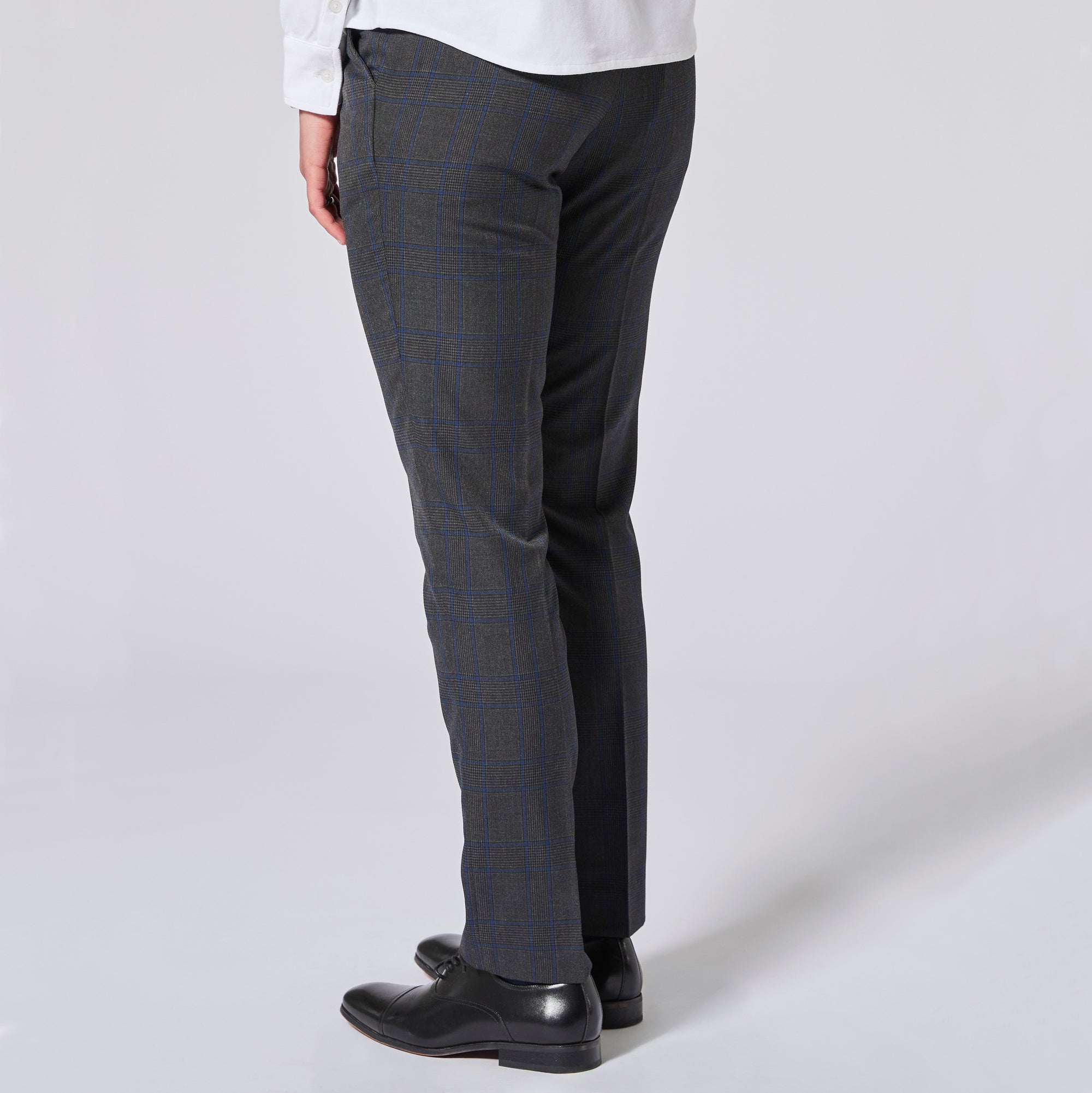 A person shown from the waist down wearing dark charcoal gray plaid trousers with a subtle blue windowpane pattern, paired with a white shirt and black leather dress shoes, against a light gray studio background. Color: Gray Admiral Plaid