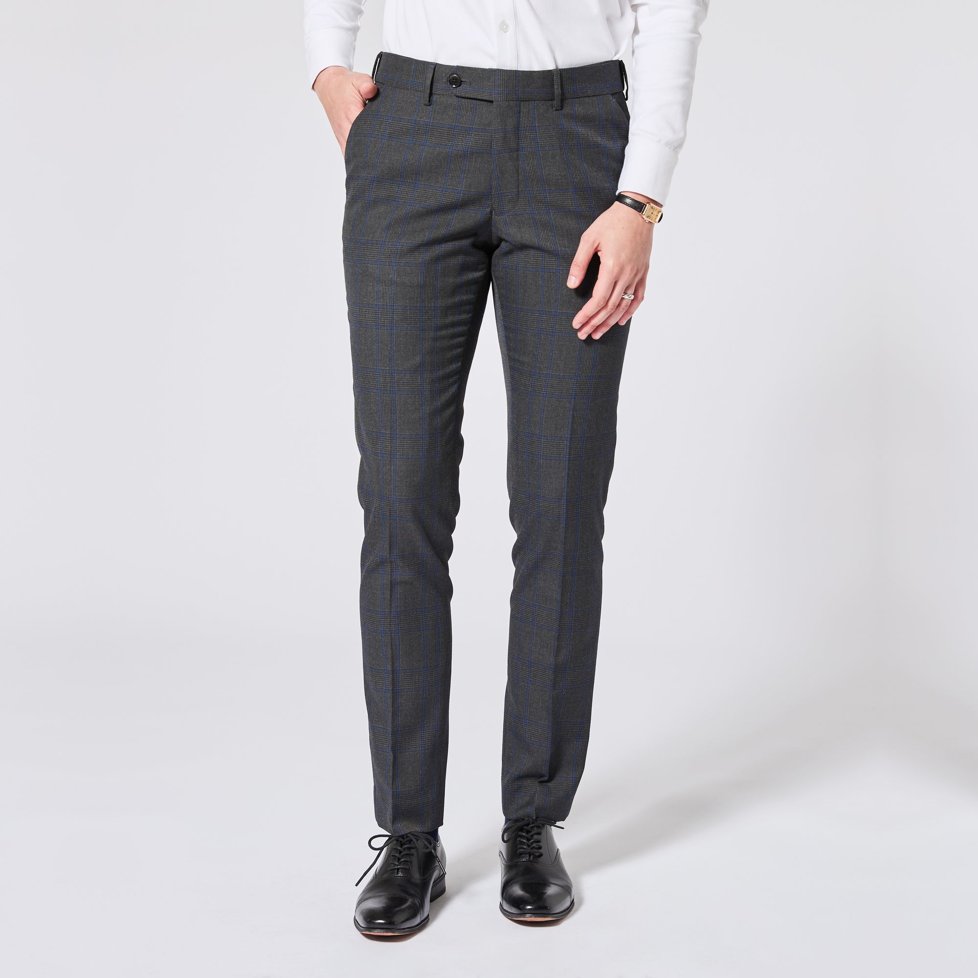 A person wearing slim-fit dark gray plaid trousers, a white long-sleeve shirt, and black leather dress shoes, standing against a light gray studio background. Color: Gray Admiral Plaid