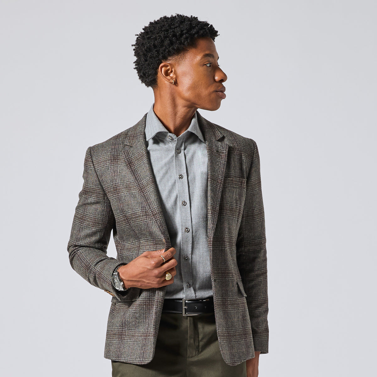 Model wearing a gray and brown plaid blazer over a light gray button-down shirt, styled with olive green trousers, against a solid light gray background. Color: Earthen Glen Plaid