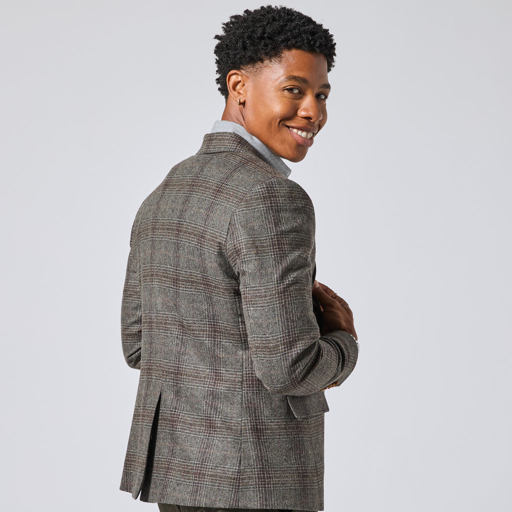 Model wearing a brown and gray plaid blazer over a light blue collared shirt, photographed from the back-three-quarter angle against a light gray studio background. Color: Earthen Glen Plaid