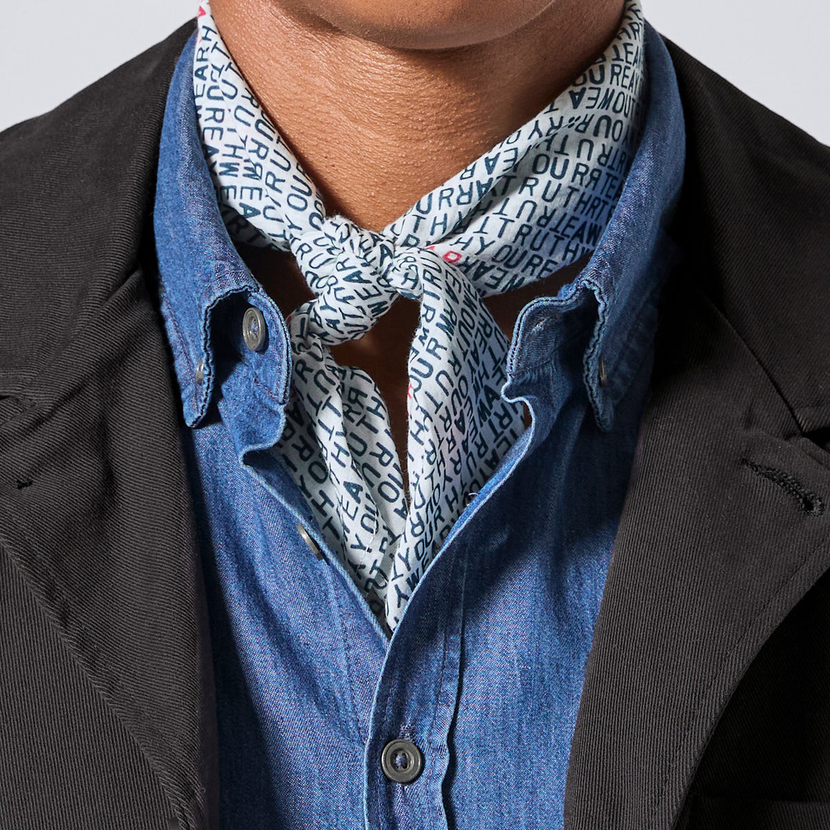 A close-up of a person wearing a light blue and white patterned bandana tied around a blue button-down shirt, layered under a black jacket.