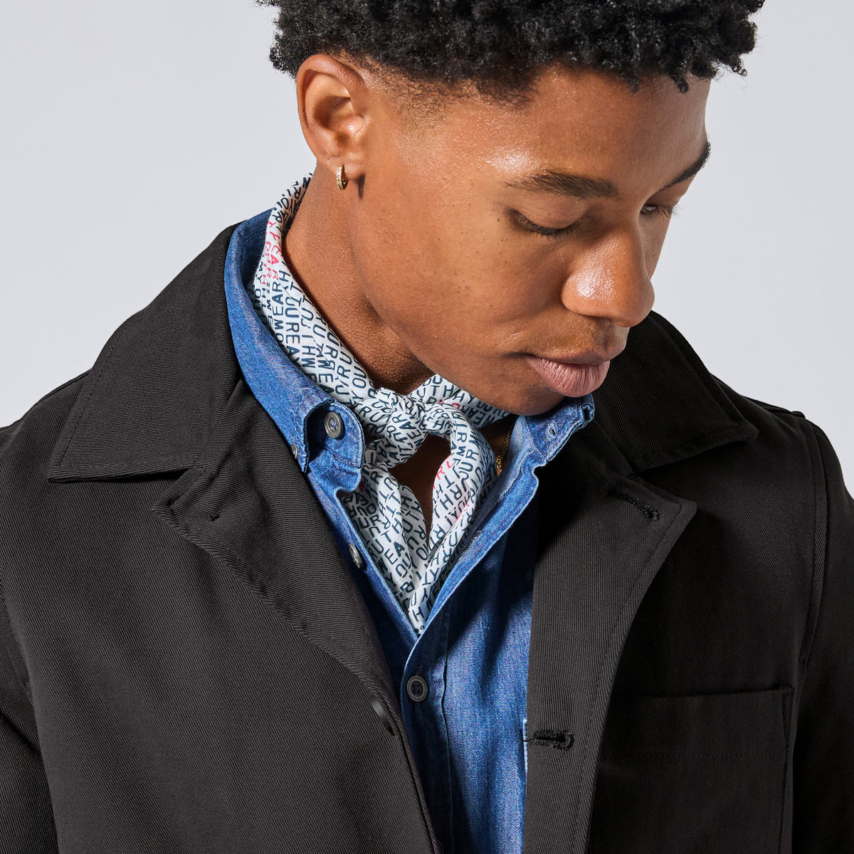 A close-up of a model wearing a black coat over a blue denim shirt and a patterned bandana tied around their neck looking down against a light gray background.