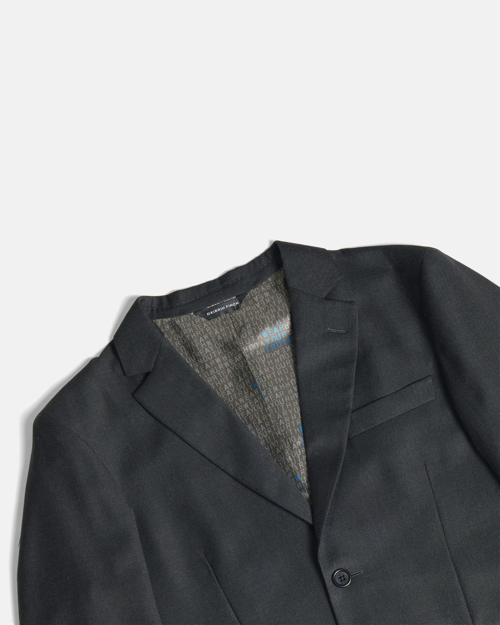 Close-up of a dark charcoal gray blazer with a textured gray and blue patterned lining visible inside the collar, set against a clean off-white background. Color: Nightfall Birdseye