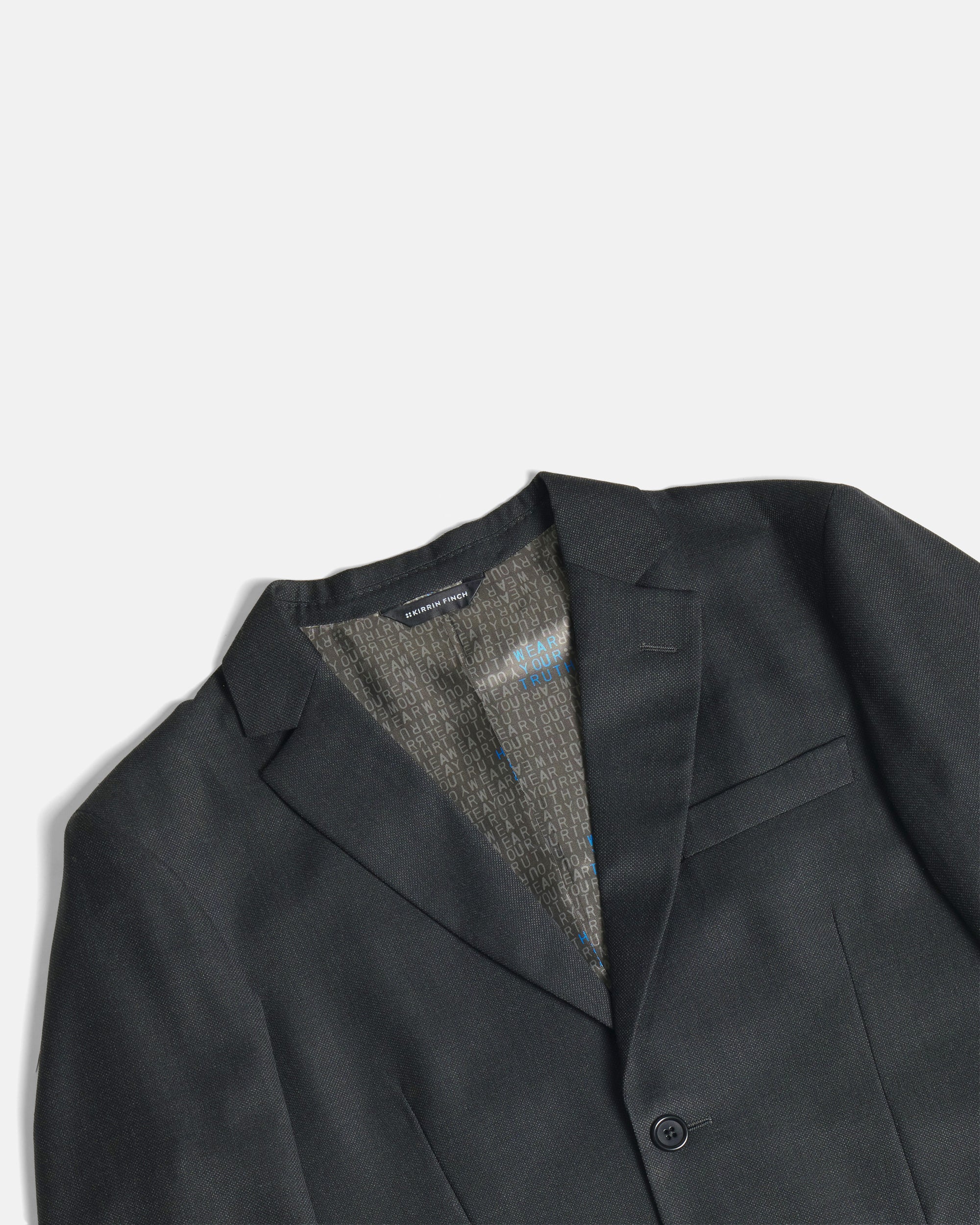 Close-up of a dark charcoal gray blazer with a textured gray and blue patterned lining visible inside the collar, set against a clean off-white background. Color: Nightfall Birdseye