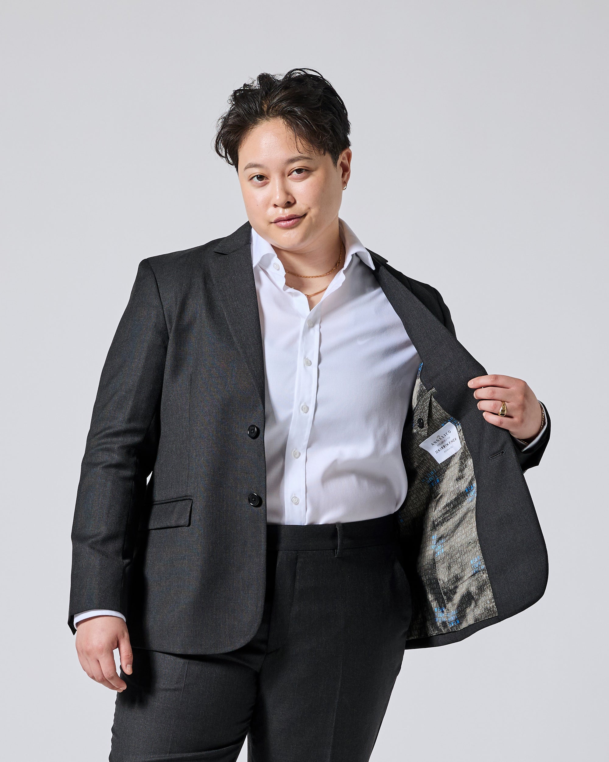 A person wearing a tailored dark gray blazer over a white button-up shirt and black trousers, standing against a light gray studio background and opening the blazer to show the interior lining. Color: Nightfall Birdseye