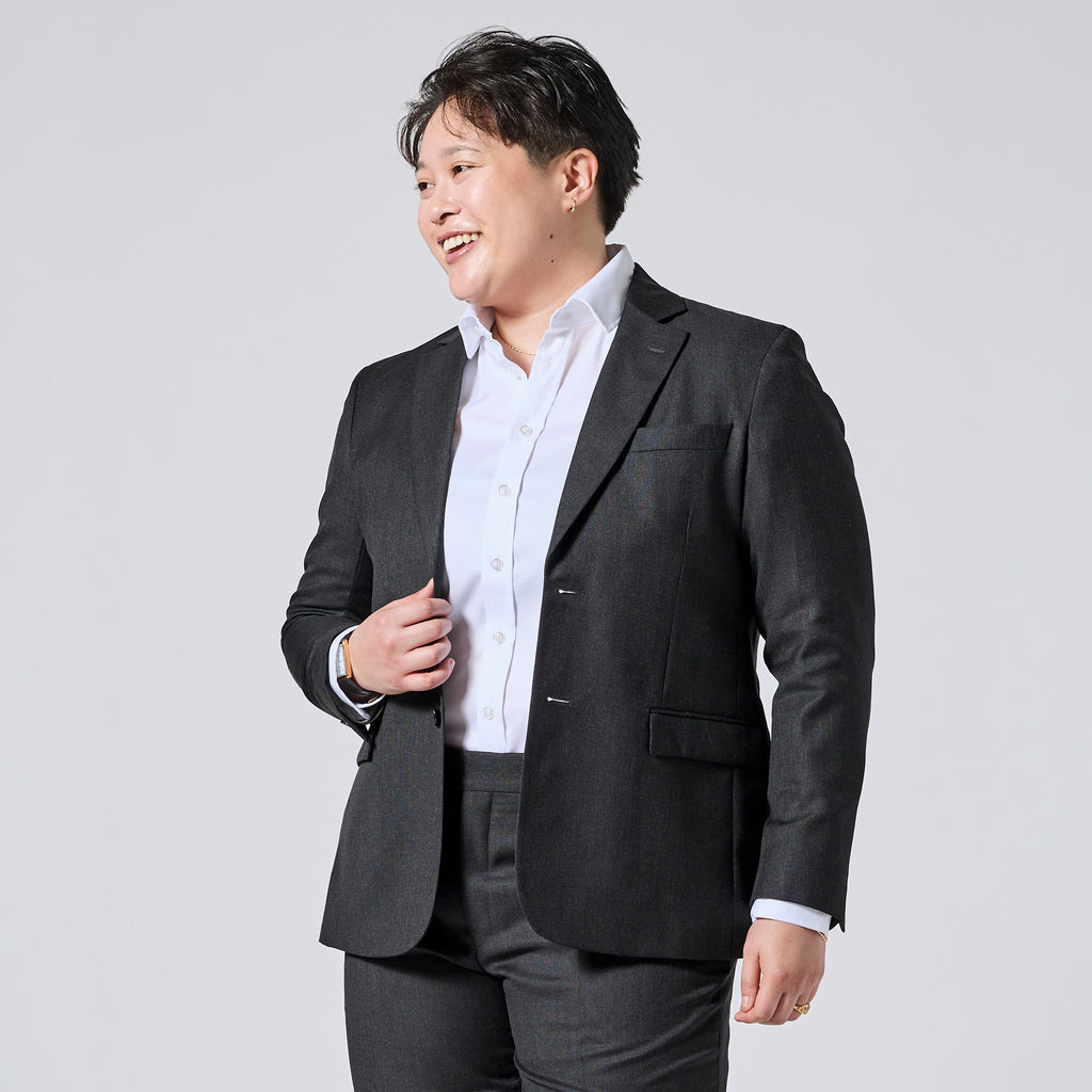 Person wearing a dark gray blazer over a white button-down shirt and dark gray trousers, smiling and looking to the side against a light gray studio background. Color: Nightfall Birdseye