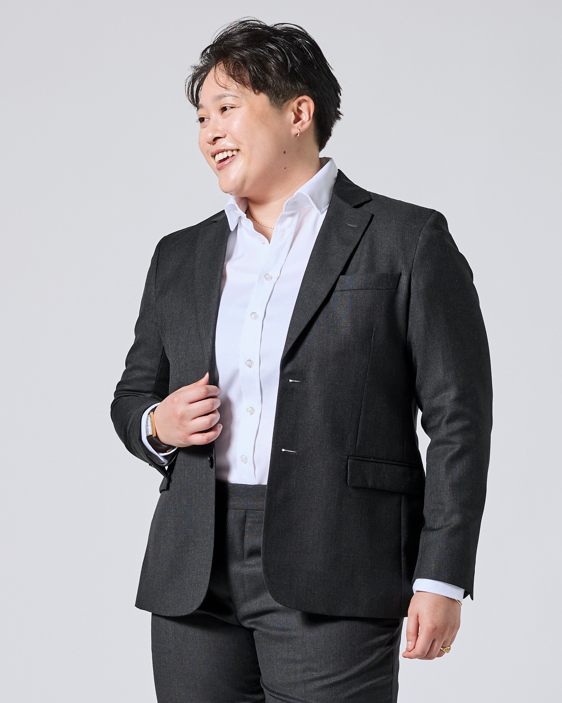 Person wearing a dark gray blazer over a white button-down shirt and dark gray trousers, smiling and looking to the side against a light gray studio background. Color: Nightfall Birdseye