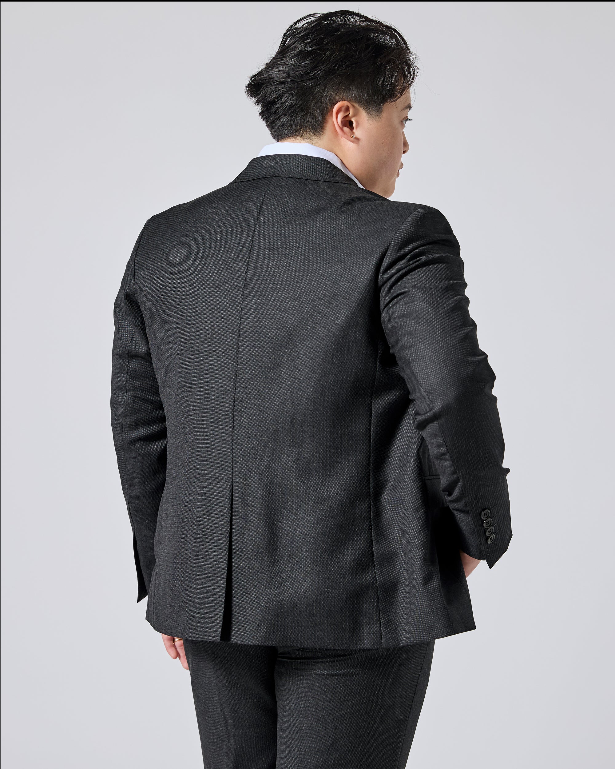 Person wearing a dark charcoal gray suit jacket, shown from the back and three-quarter side view against a light gray studio background. Color: Nightfall Birdseye