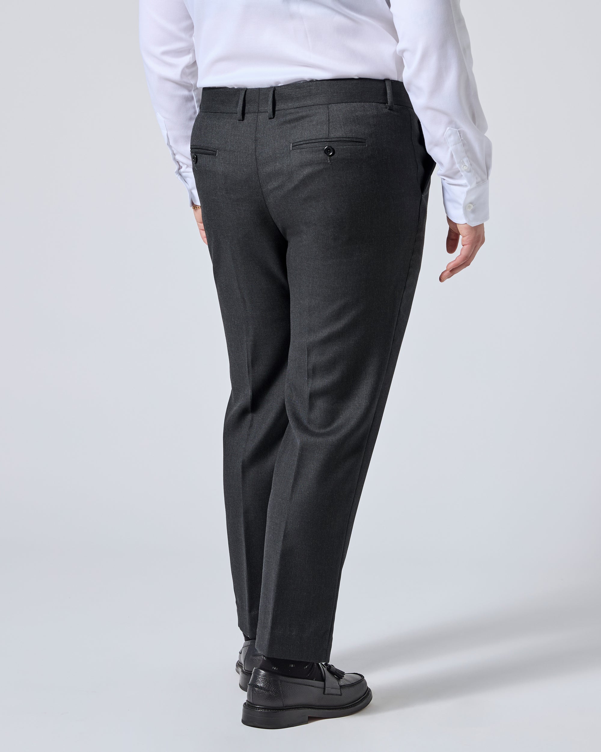 A person shown from the back wearing dark charcoal gray dress pants, a white long-sleeve shirt, and black loafers against a light gray studio background. Color: Nightfall Birdseye