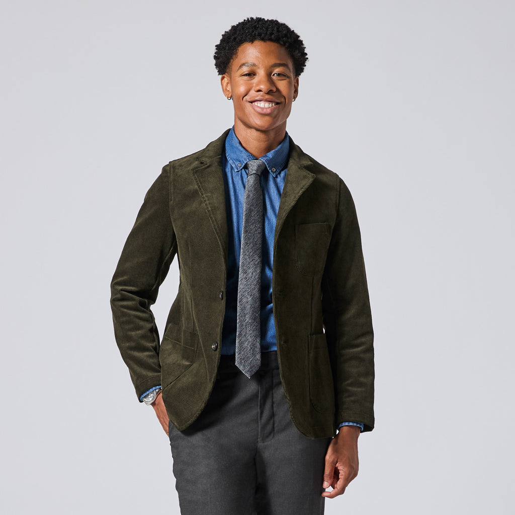Model wearing an olive green corduroy blazer over a blue button-down shirt and a gray patterned tie, paired with dark gray trousers, photographed against a solid light gray studio background. Color: Pine