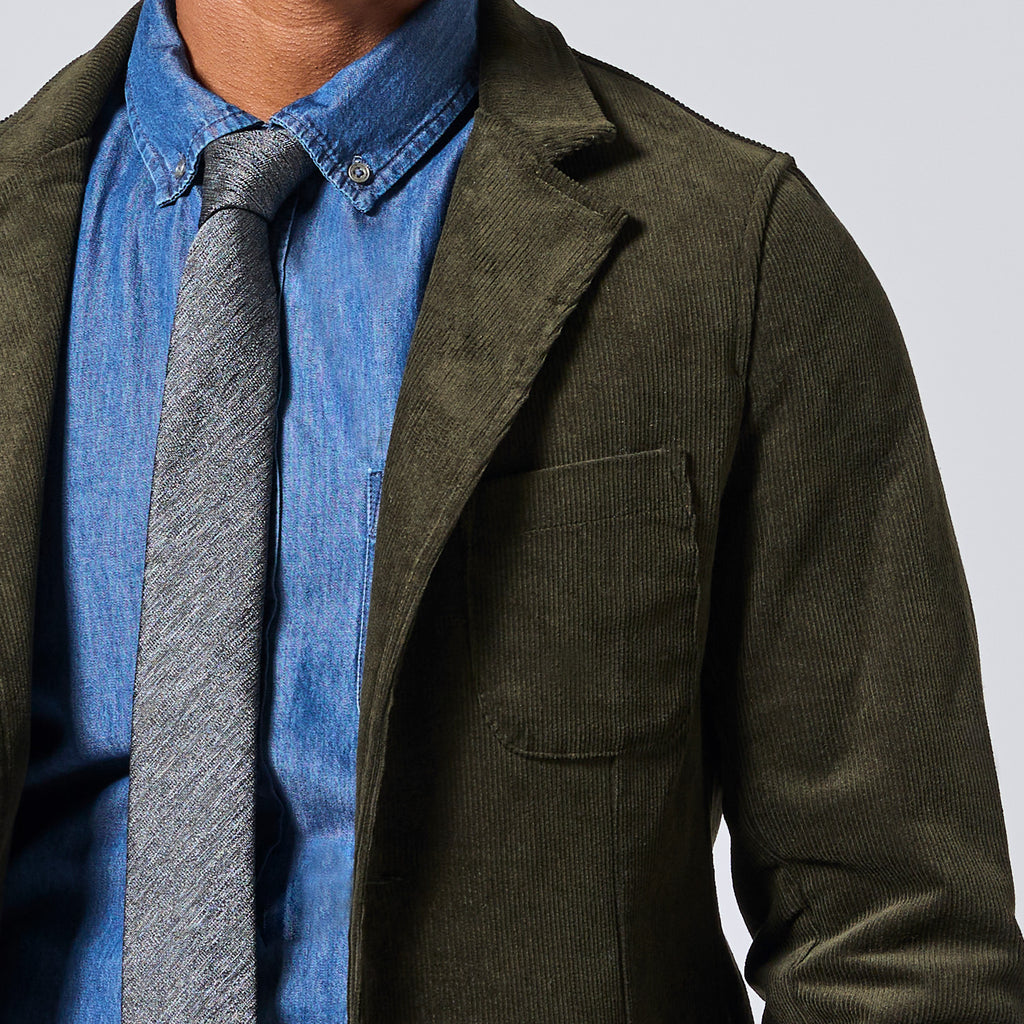 Close-up of a person wearing an olive green corduroy blazer over a blue button-down shirt with a gray textured tie, against a light gray background. Color: Pine