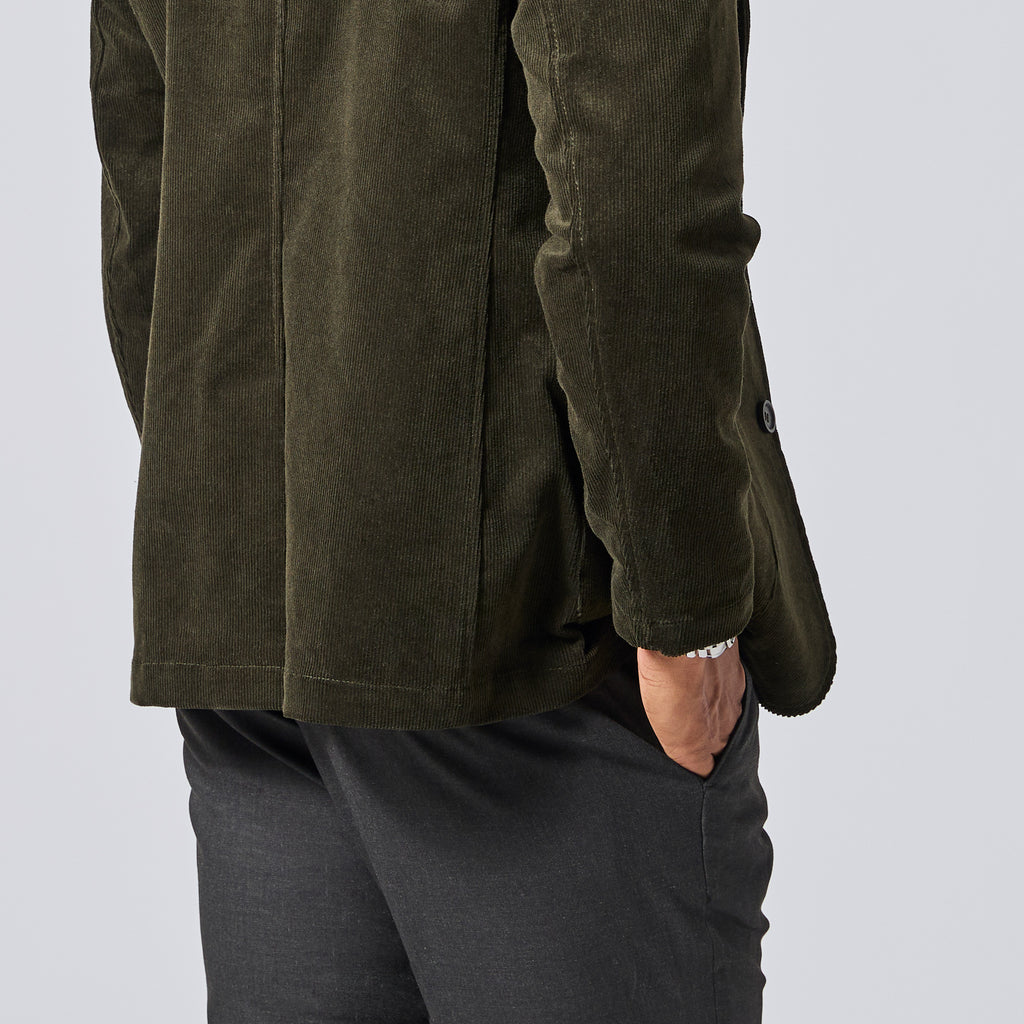 Close-up of a person wearing an olive green corduroy blazer with dark gray trousers, standing against a light gray studio background. Color: Pine