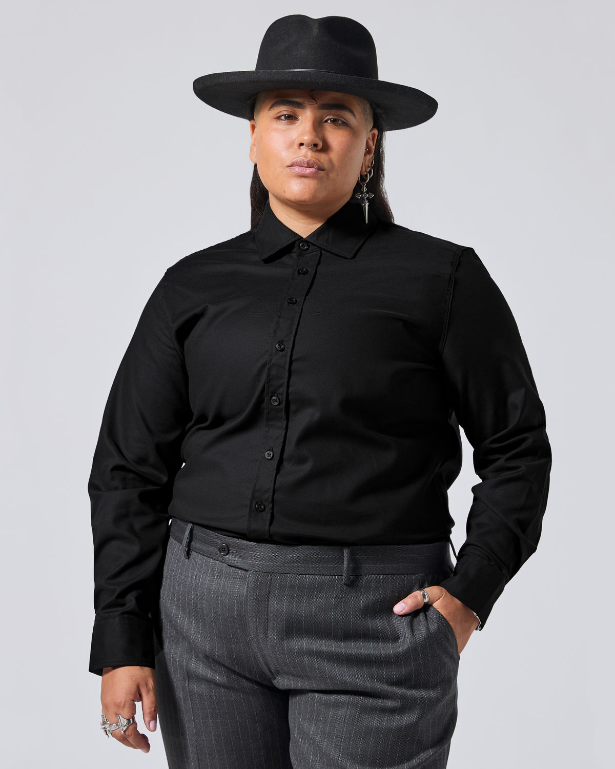 A model wearing a black long-sleeve button-down shirt, a black wide-brim fedora hat, and dark gray pinstripe trousers, posed against a solid light gray background. Color: Black
