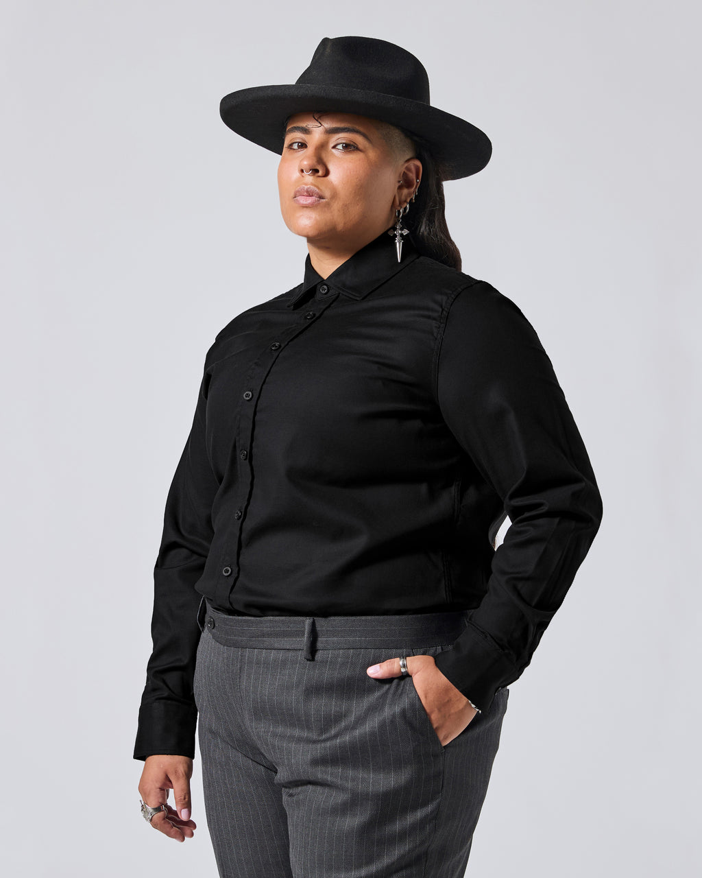 A model wearing a black long-sleeve button-up shirt, a black wide-brim hat, and gray pinstripe trousers, photographed against a solid light gray background. Color: Black