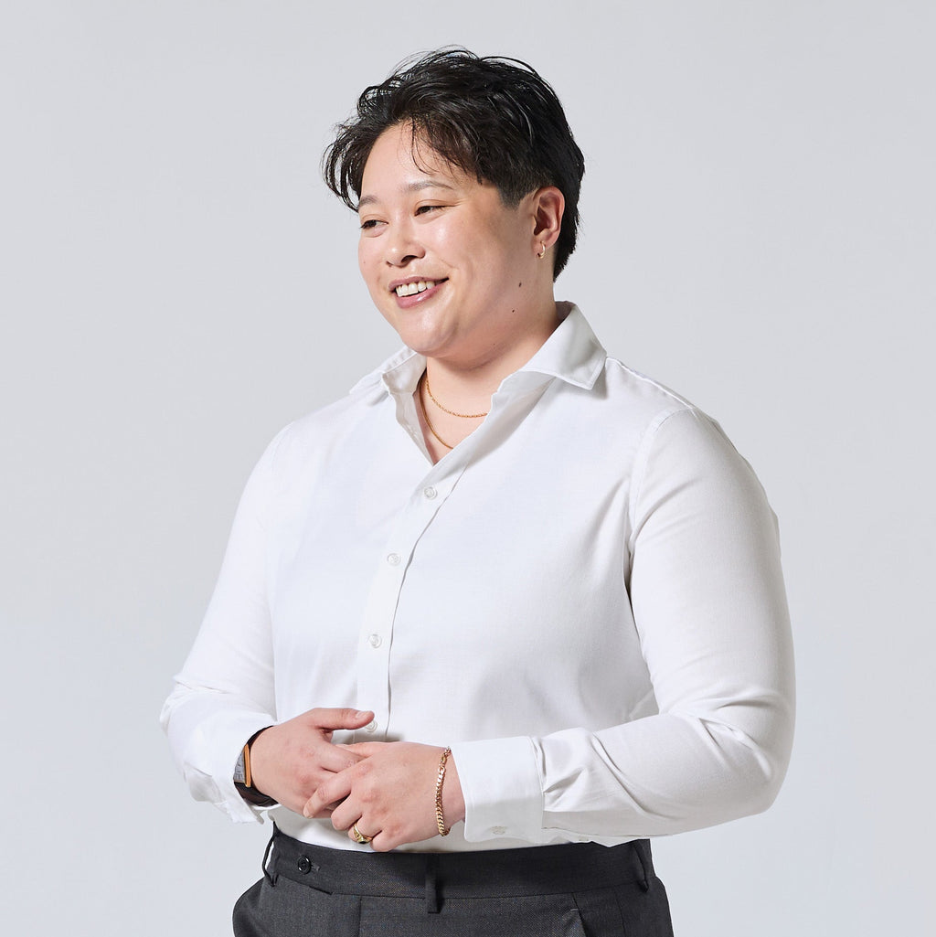 Model wearing a white long-sleeve button-down shirt and dark gray trousers, standing against a light gray studio background. Color: White