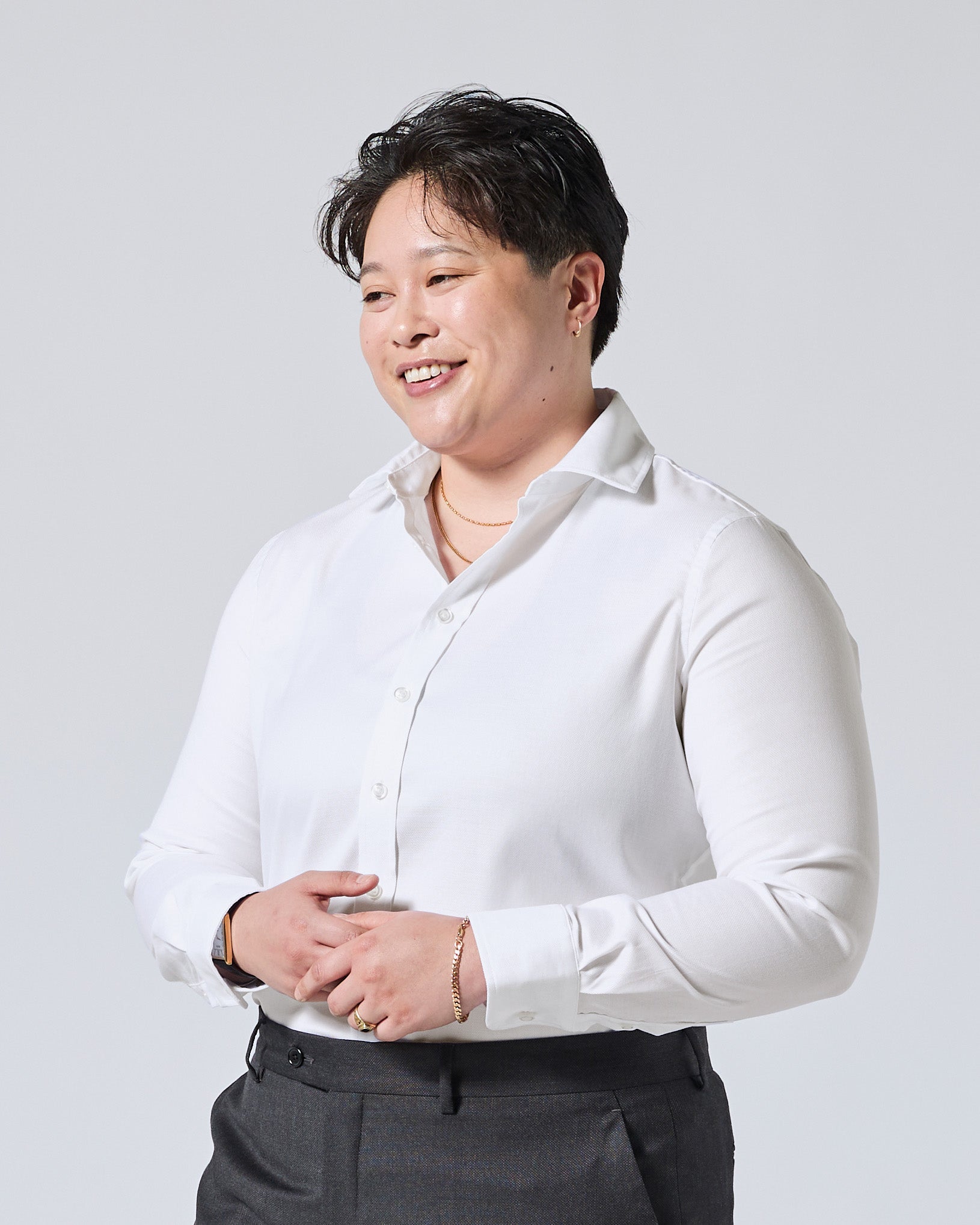Model wearing a white long-sleeve button-down shirt and dark gray trousers, standing against a light gray studio background. Color: White