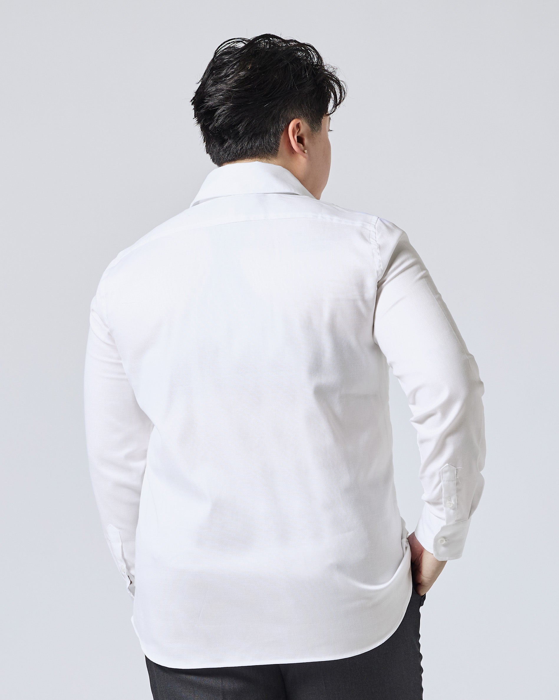 Model viewed from behind wearing a white long-sleeve button-down shirt and dark trousers, standing against a light gray studio background. Color: White