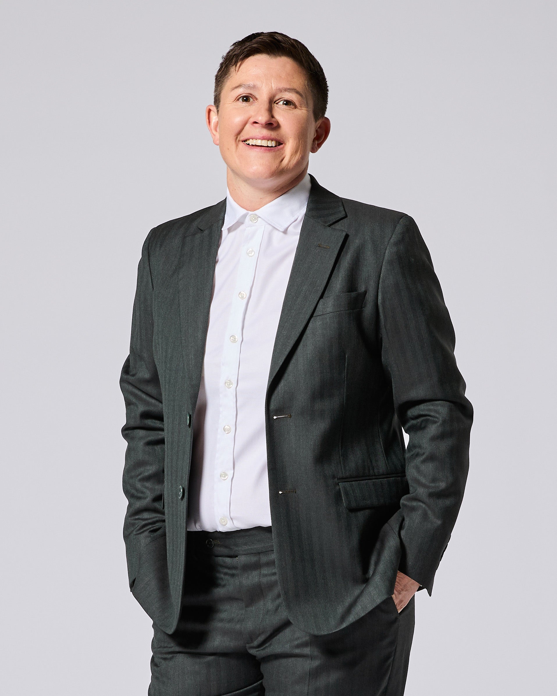 A smiling person wearing a dark gray pinstripe blazer over a white button-down shirt, paired with matching gray trousers, photographed against a solid light gray studio background. Color: Midnight Pine Herringbone