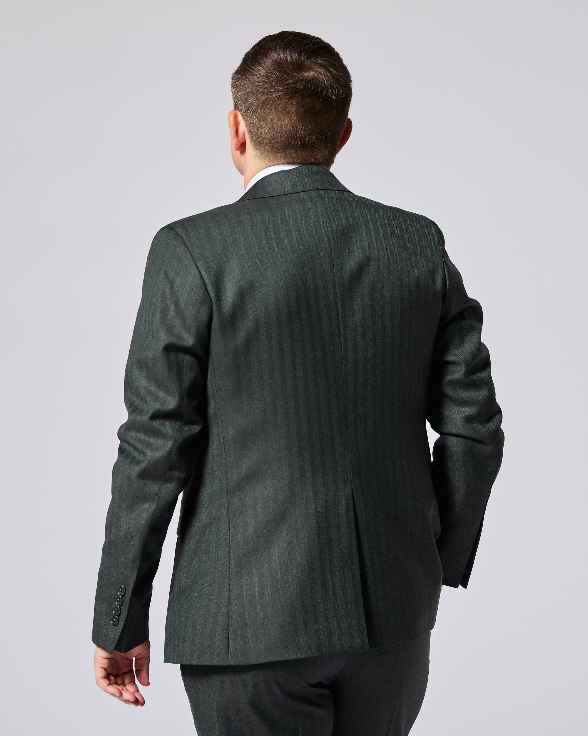 Person viewed from the back wearing a dark green suit jacket and matching trousers against a light gray studio background. Color: Midnight Pine Herringbone
