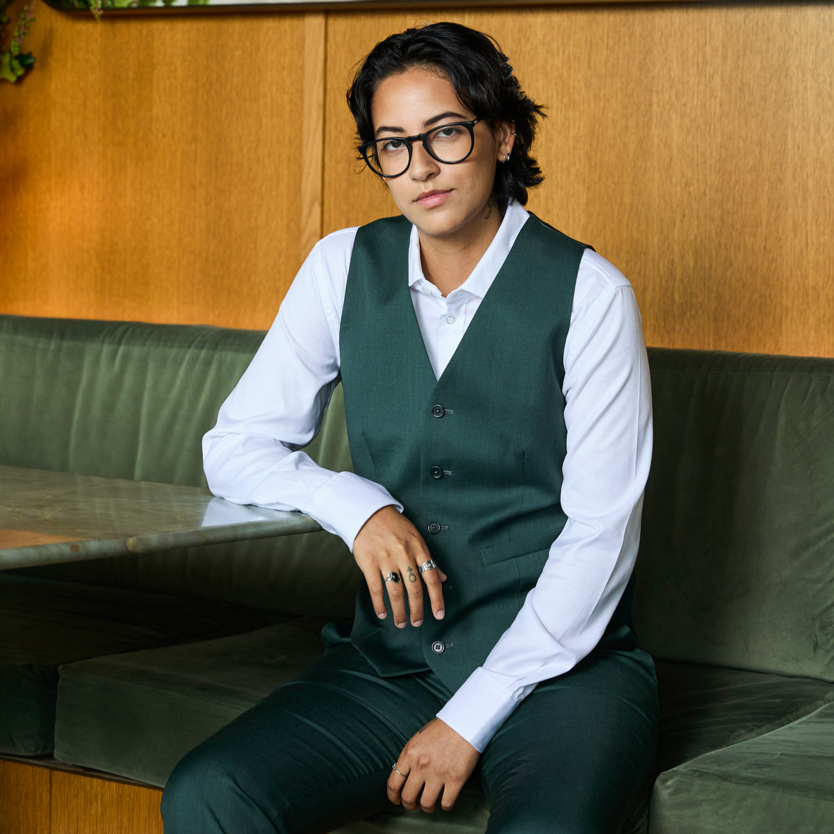 A person wearing a dark green vest and matching pants over a white long-sleeve shirt, seated on a green bench in a restaurant or cafe setting with wood paneling in the background. Color: Emerald