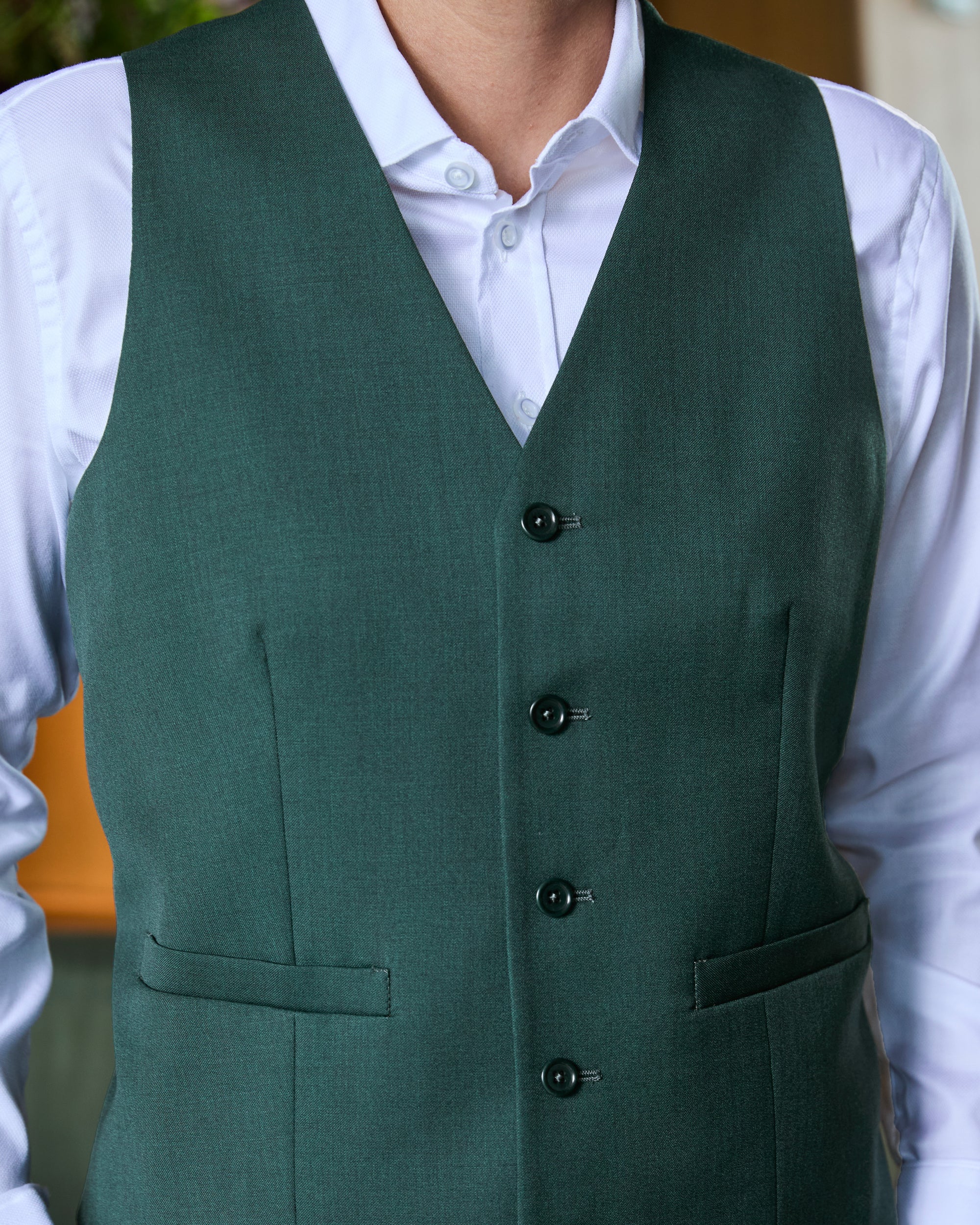 A dark green tailored waistcoat vest worn over a white collared dress shirt, shown from the chest down on a person. Color: Emerald