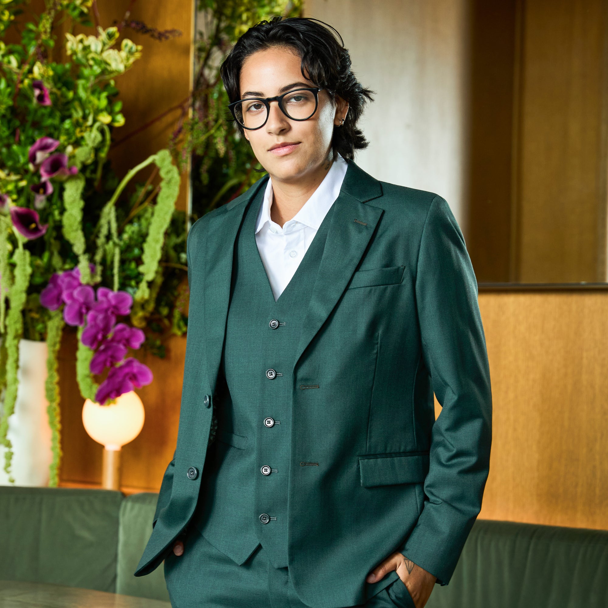 Model wearing a dark green three-piece suit with a white collared shirt, standing in a warm indoor setting with a floral arrangement in the background. Color: Emerald