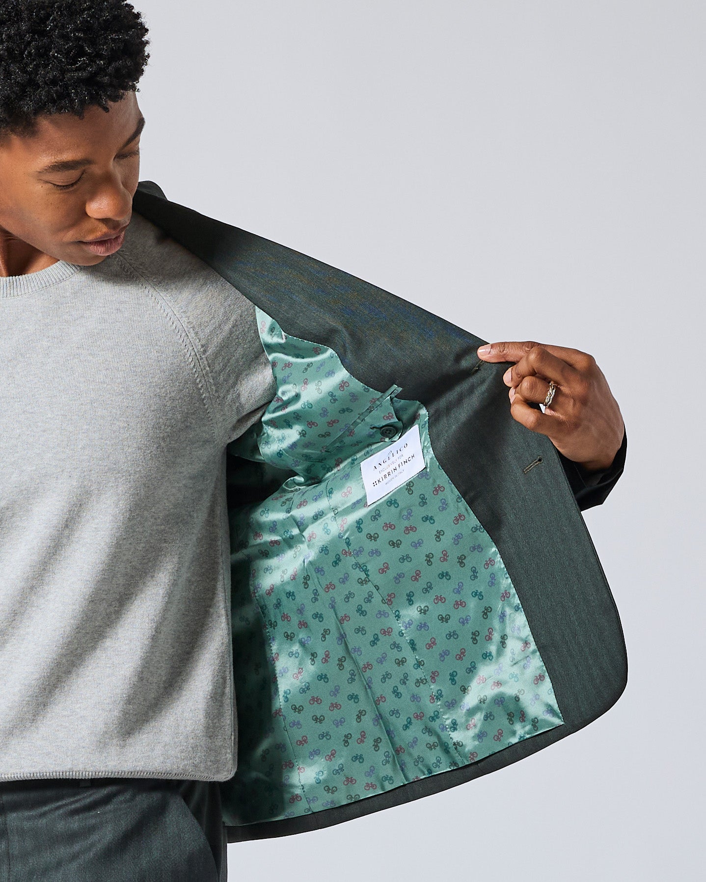 Person wearing a gray sweater and dark trousers holds open a dark green suit jacket to show the interior lining.  Color: Midnight Pine Herringbone