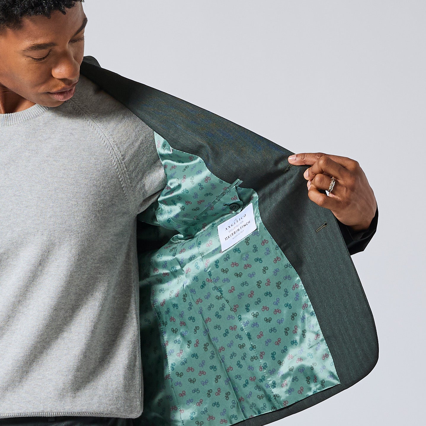 A close-up of a person wearing a gray sweater and dark green blazer, opening the blazer to show a green satin-lined interior. Color: Midnight Pine Herringbone