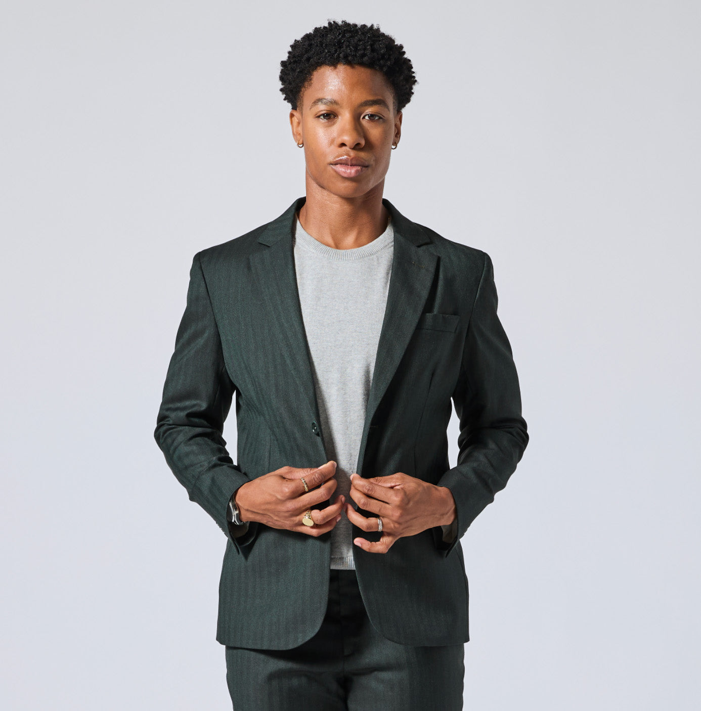A man wearing a dark green pinstripe blazer over a light gray crewneck t-shirt, paired with matching dark green trousers, photographed against a solid light gray background. Color: Midnight Pine Herringbone