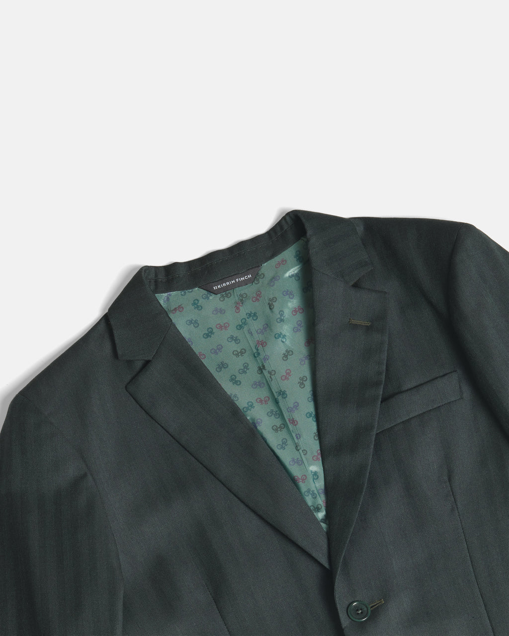 A close-up of a dark green suit jacket, showing the textured fabric, notch lapels, and a patterned green inner lining. Color: Midnight Pine Herringbone