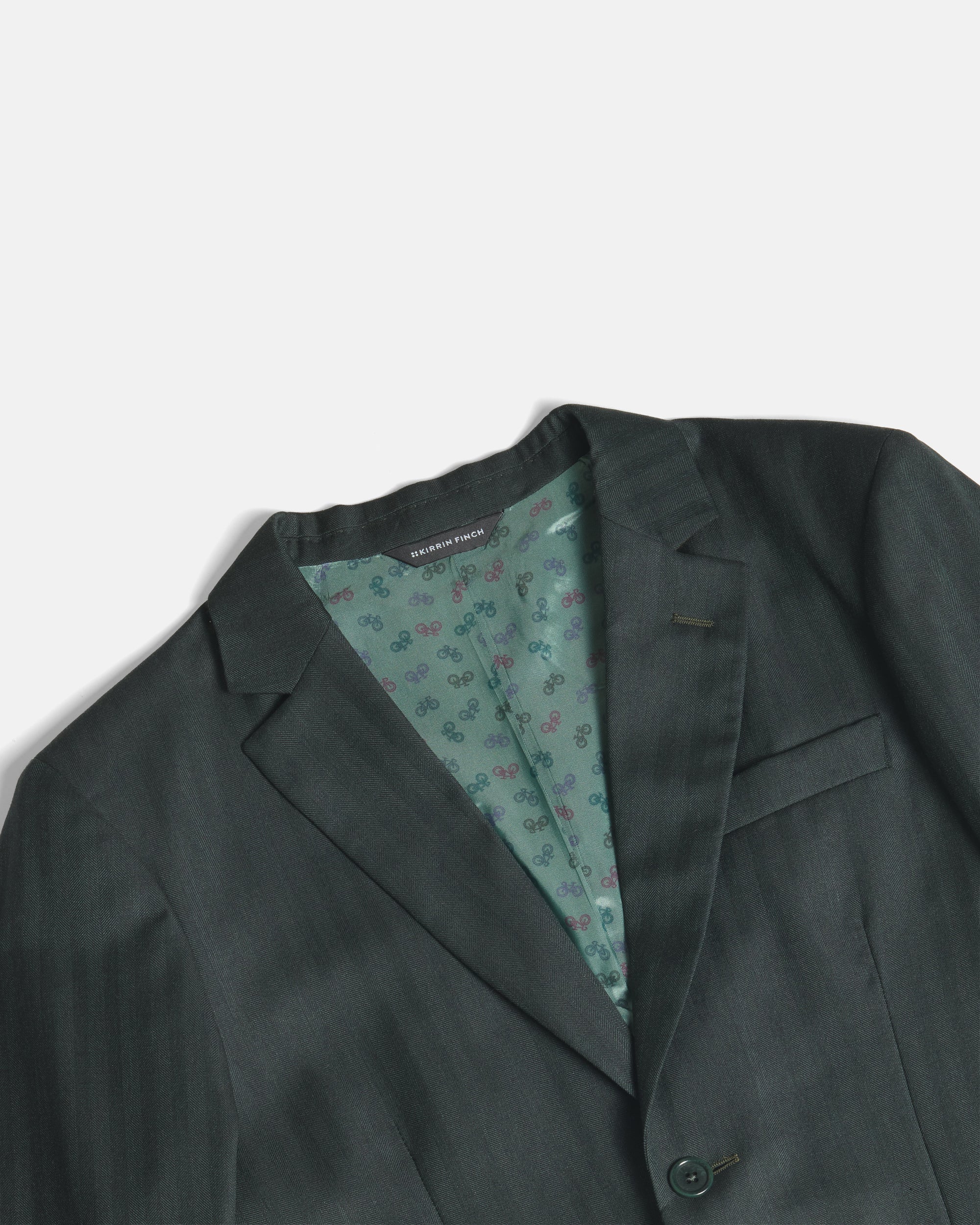 A close-up of a dark green suit jacket, showing the textured fabric, notch lapels, and a patterned green inner lining. Color: Midnight Pine Herringbone