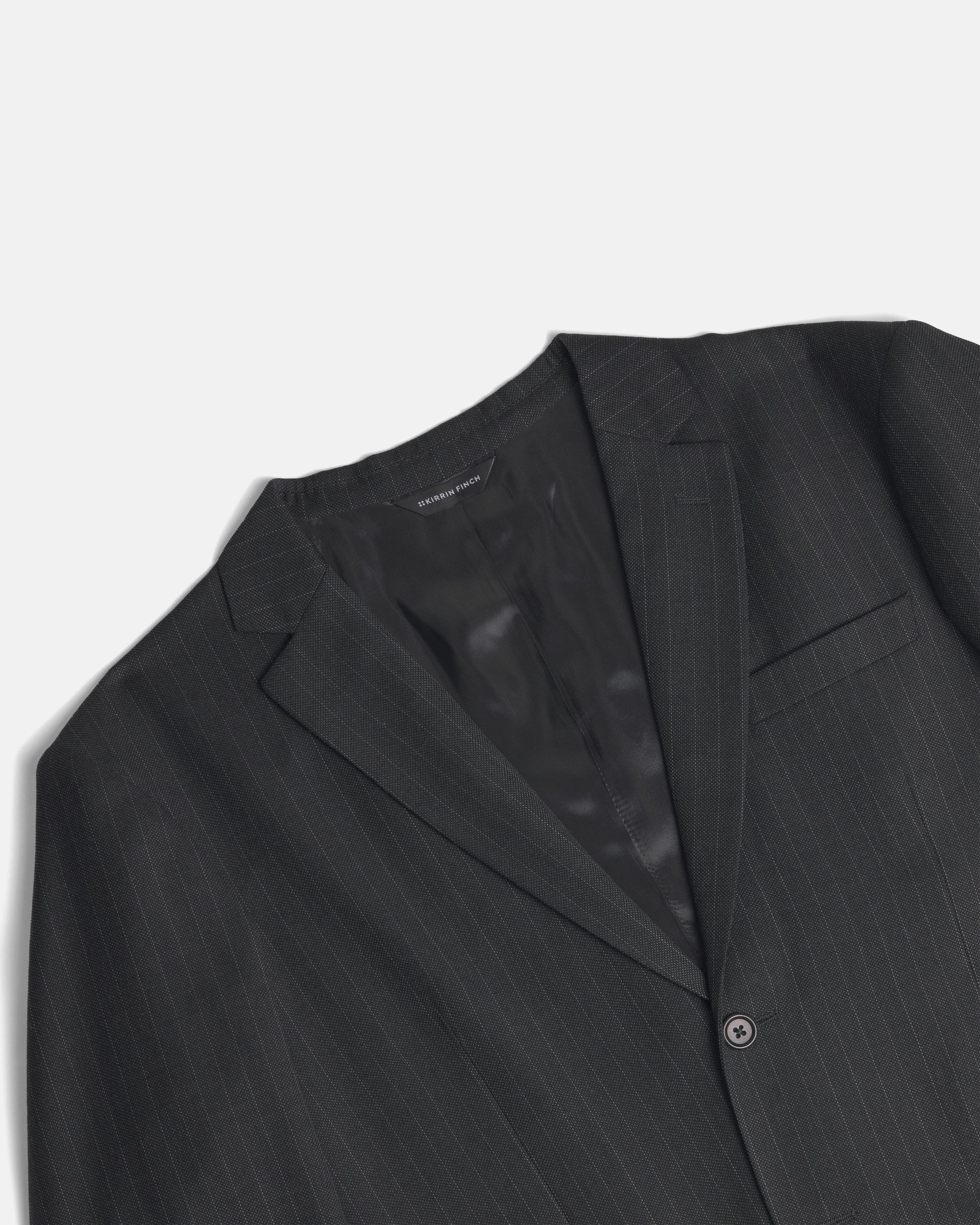 A close-up of a dark charcoal gray pinstripe suit jacket, showing the lapel, inner collar, and a single button closure against a light gray background. Color: Shadow Pinstripe