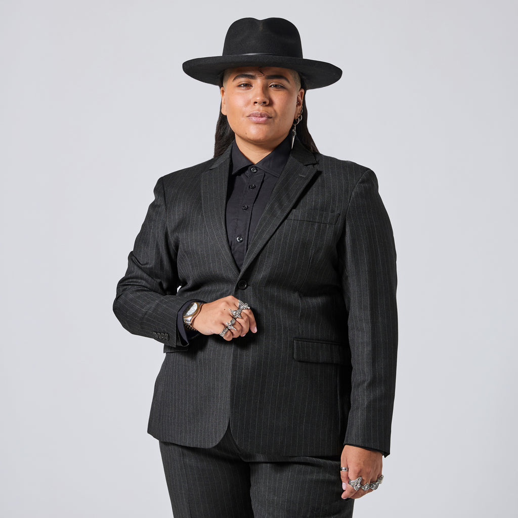 A model wearing a black pinstripe blazer, matching trousers, a black button-down shirt, and a black wide-brim hat, posed against a light gray studio background. Color: Shadow Pinstripe