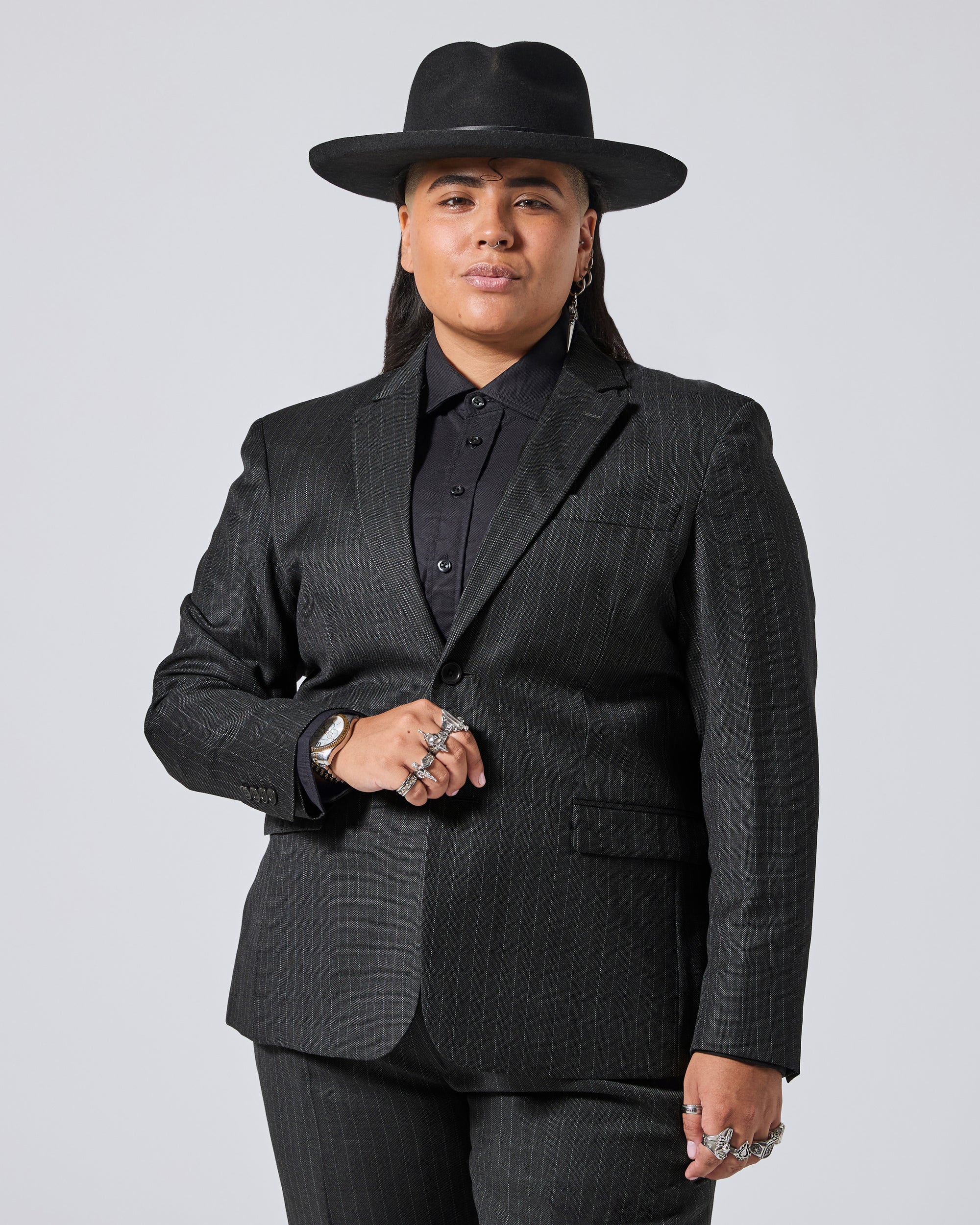 A model wearing a black pinstripe blazer, matching trousers, a black button-down shirt, and a black wide-brim hat, posed against a light gray studio background. Color: Shadow Pinstripe