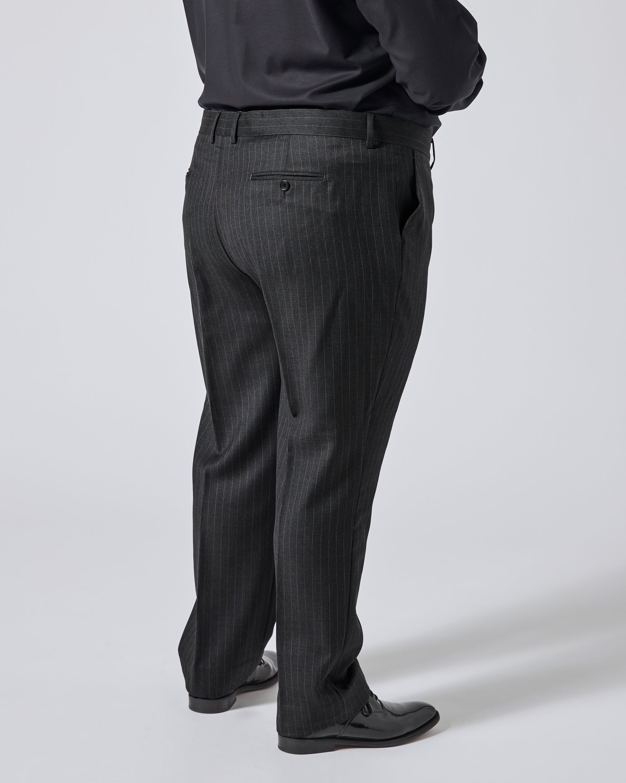 A person shown from the back wearing dark charcoal pinstripe trousers, a black shirt, and black dress shoes against a light gray studio background. Color: Shadow Pinstripe