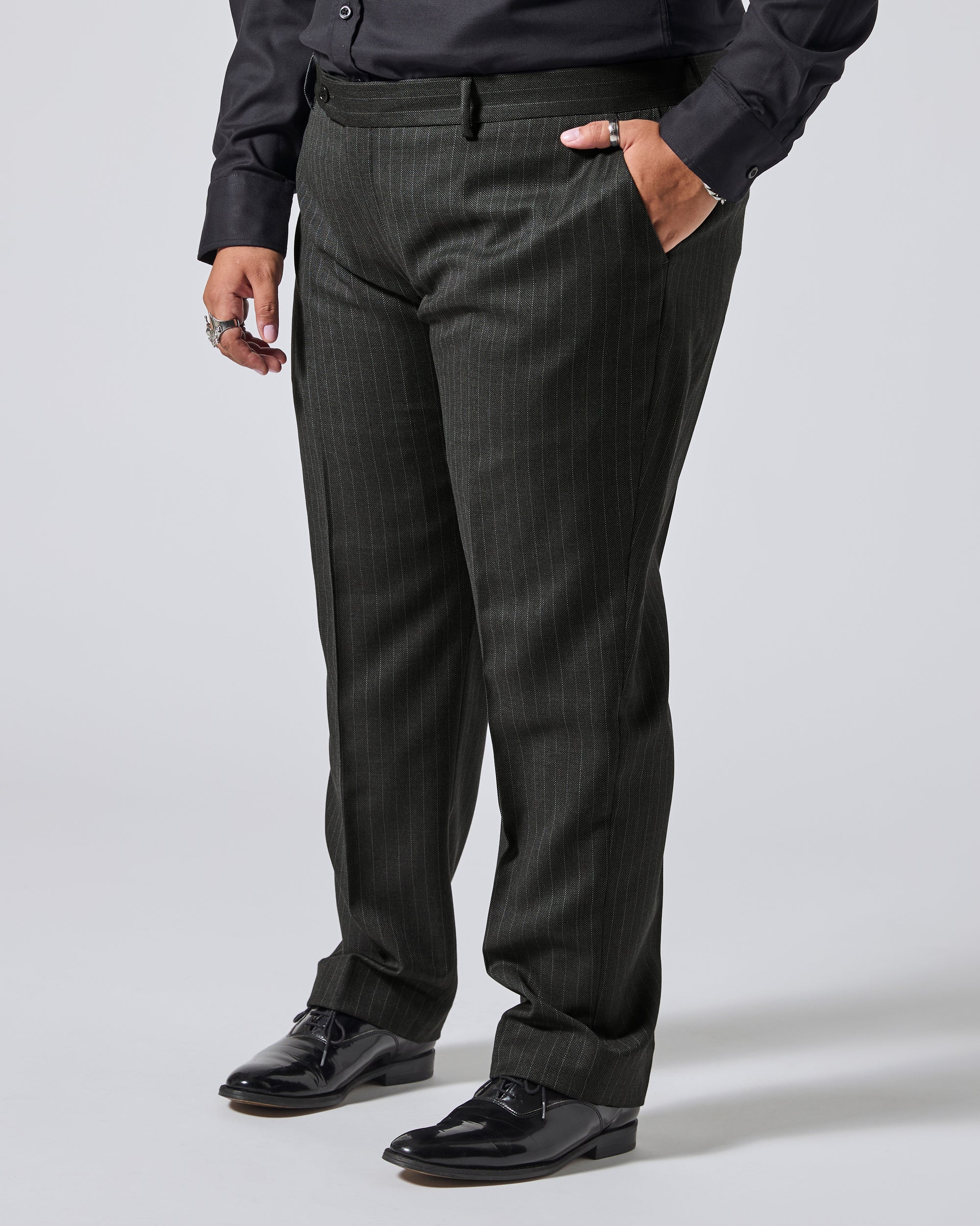 A person wearing dark charcoal gray pinstriped dress pants with a black shirt and black dress shoes, standing against a light gray studio background. Color: Shadow Pinstripe